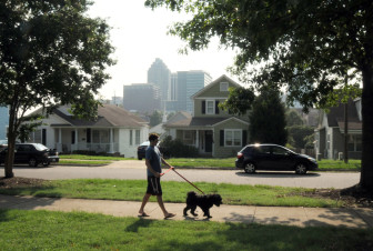 A man walks his dog down Chavis Street with a view of downtown Raleigh behind him.